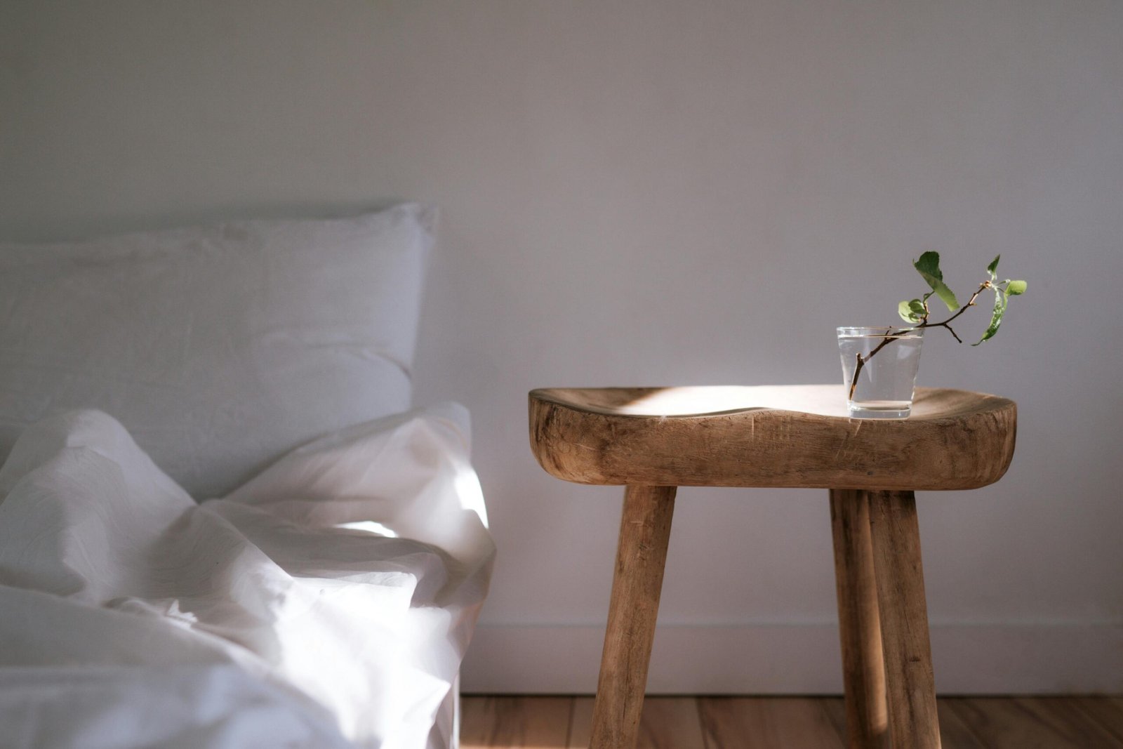 Sunlit minimalist bedroom featuring white linens and a wooden side table with a plant.